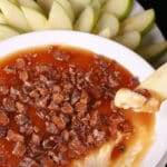 A bowl of cream cheese caramel apple dip, with caramel sauce and chopped Skor bars on top, and sliced granny smith apples behind it.