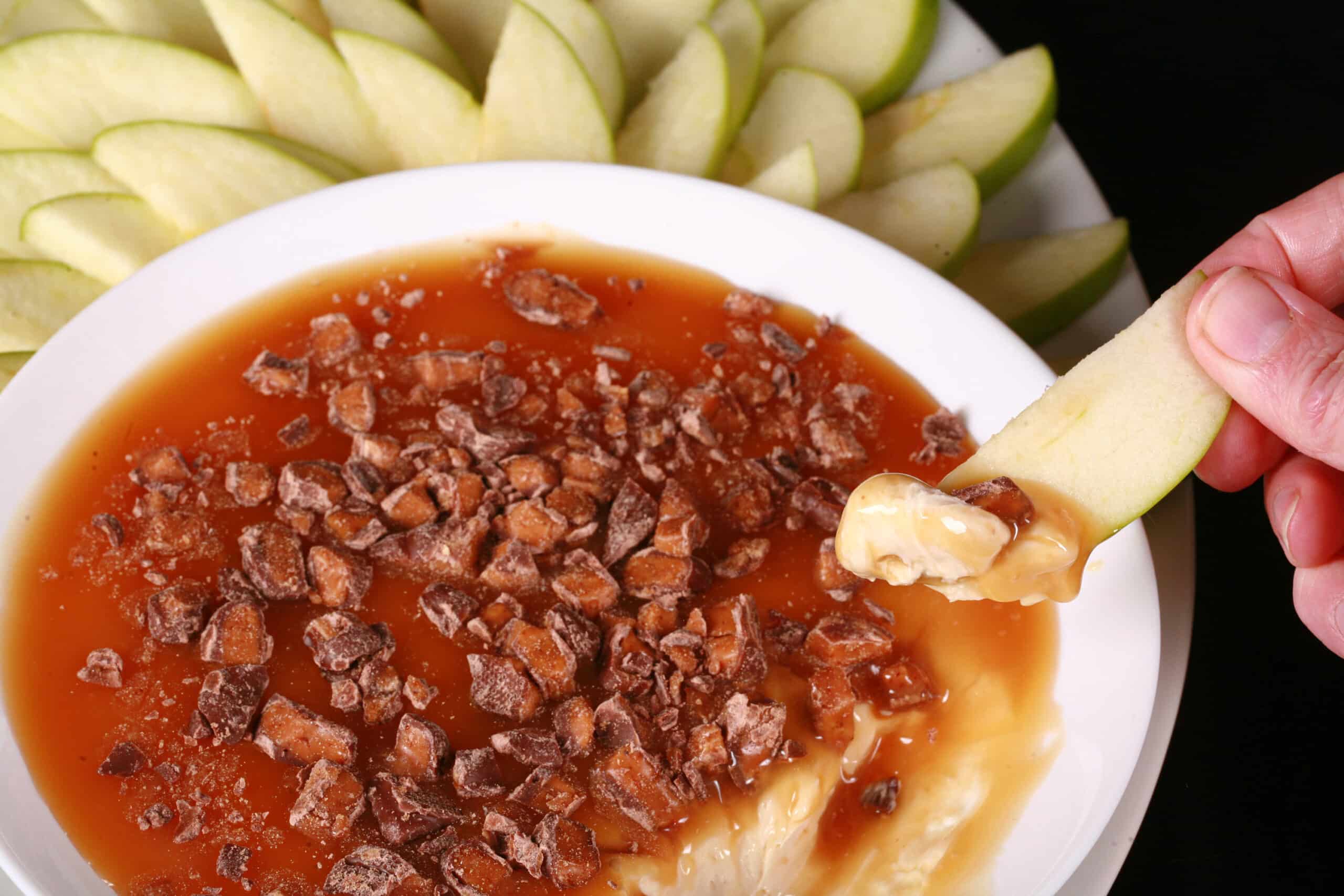 A bowl of cream cheese caramel apple dip, with caramel sauce and chopped Skor bars on top, and sliced granny smith apples behind it.