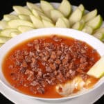 A bowl of caramel apple cheesecake dip, with caramel sauce and chopped Skor bars on top, and sliced granny smith apples behind it.