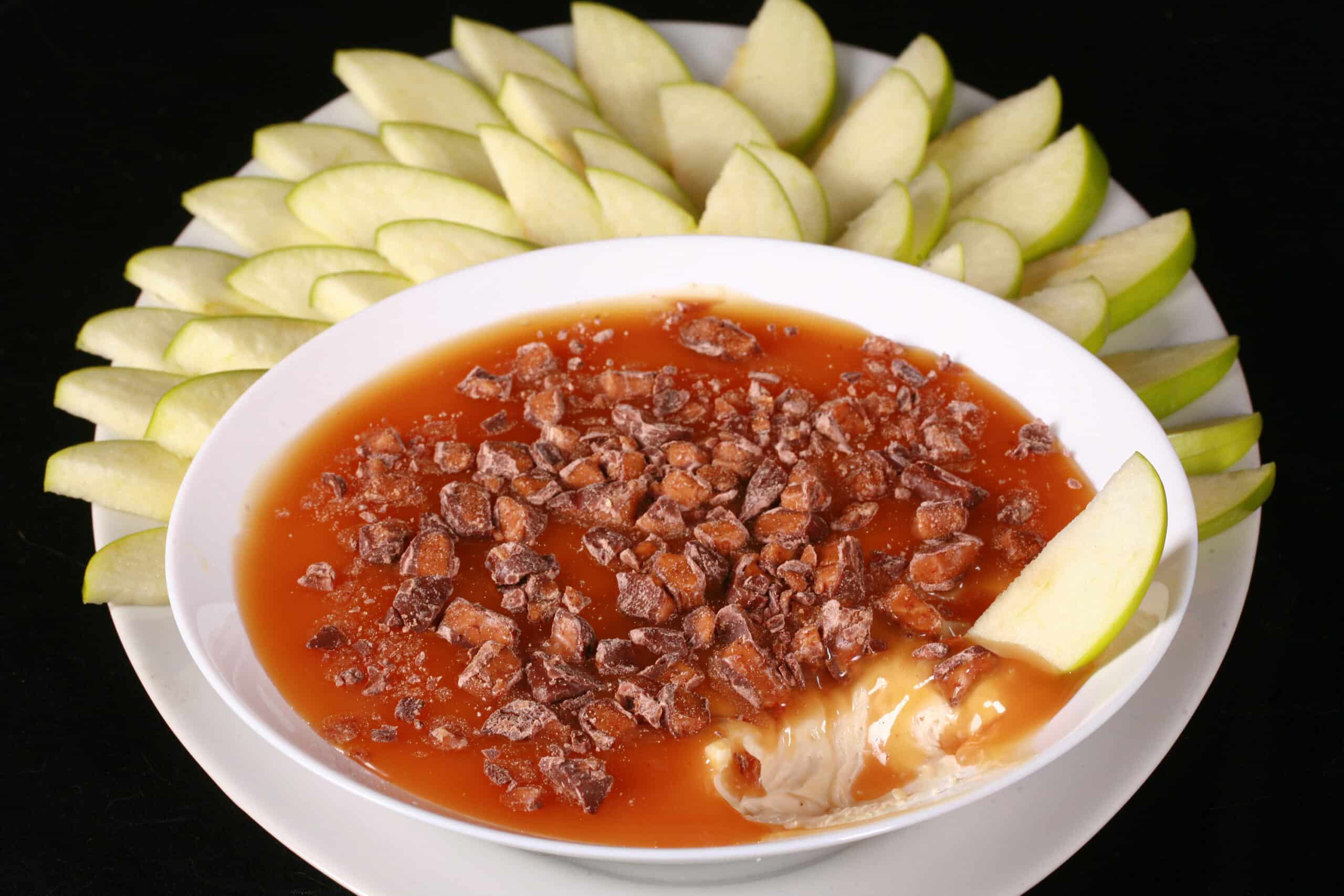 A bowl of caramel apple cheesecake dip, with caramel sauce and chopped Skor bars on top, and sliced granny smith apples behind it.