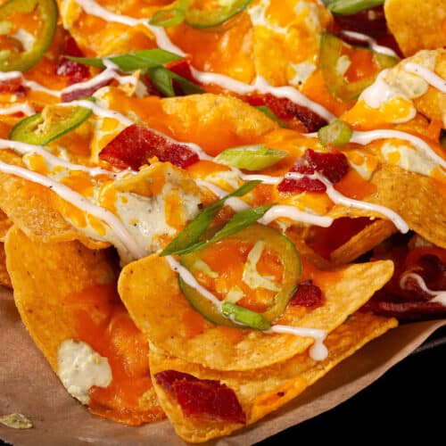 A pan of jalapeno popper nachos with jalapeno cream cheese queso, jalapeno slices, bacon, cheddar, green onions, ranch dressing, and ranch flavoured sour cream.
