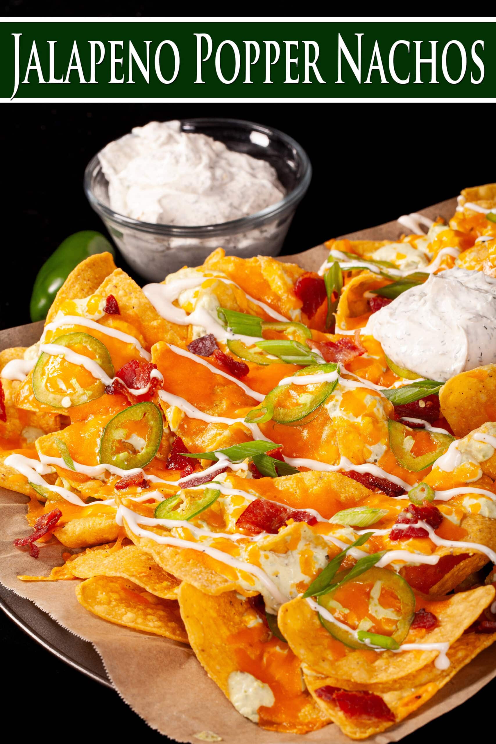 A pan of jalapeno popper nachos with jalapeno cream cheese queso,  jalapeno slices, bacon, cheddar, green onions, ranch dressing, and ranch flavoured sour cream.  Overlaid text says jalapeno popper nachos.