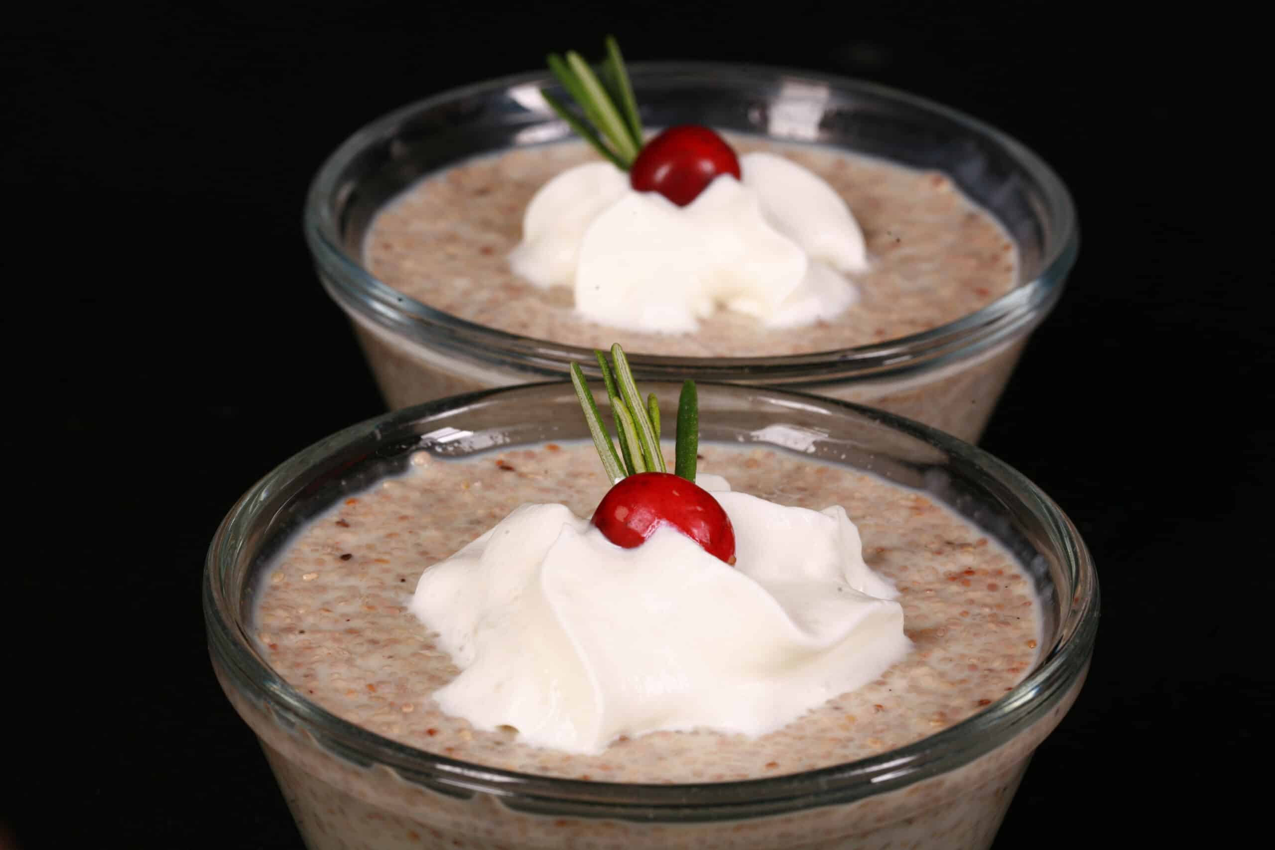 2 glass bowls of eggnog chia pudding garnished with whipped cream, a cranberry, and a sprig of rosemary.