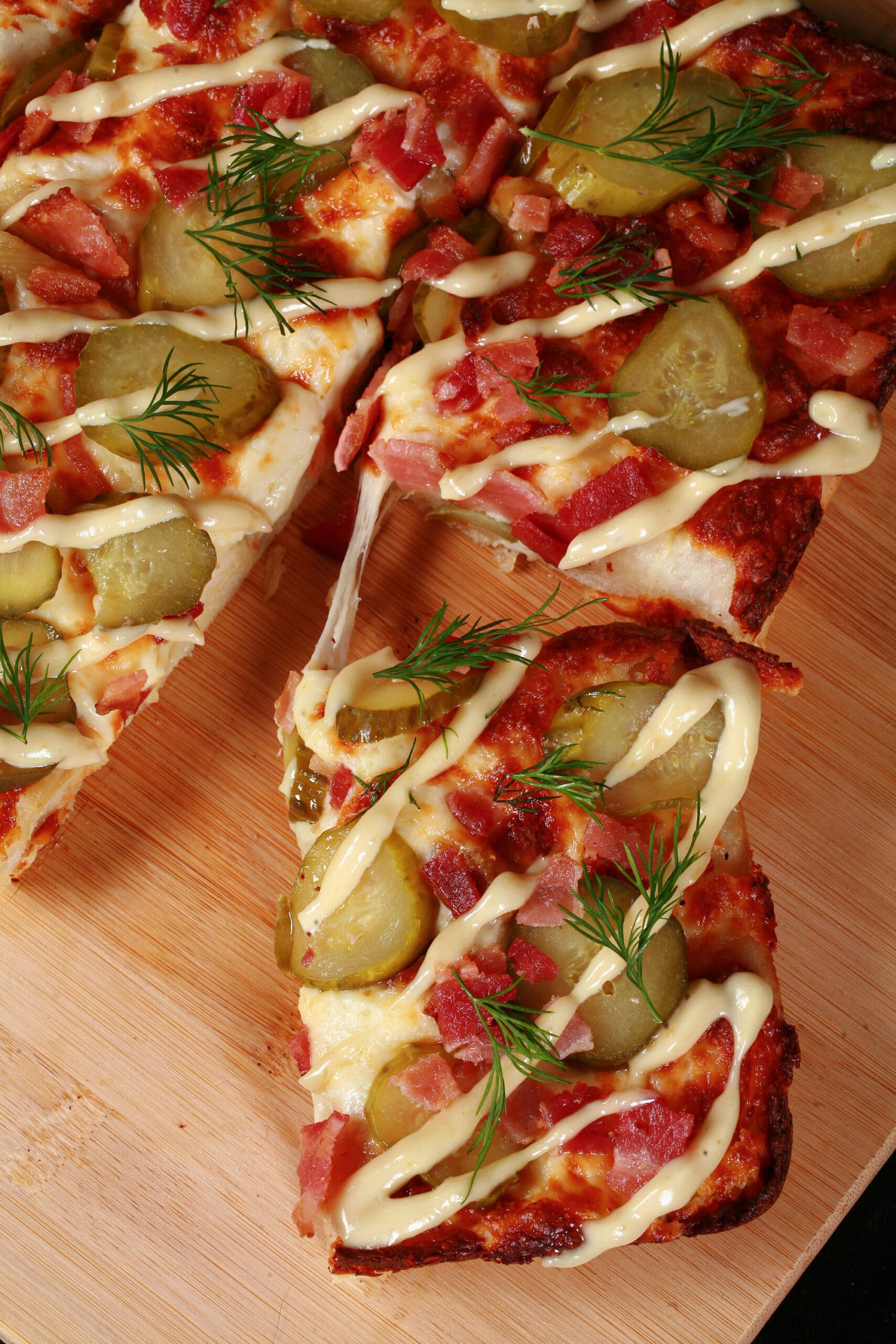 A detroit style gluten free dill pickle pizza, with pickle slices, bacon, fresh dill, and roasted garlic mayonnaise drizzled across it.