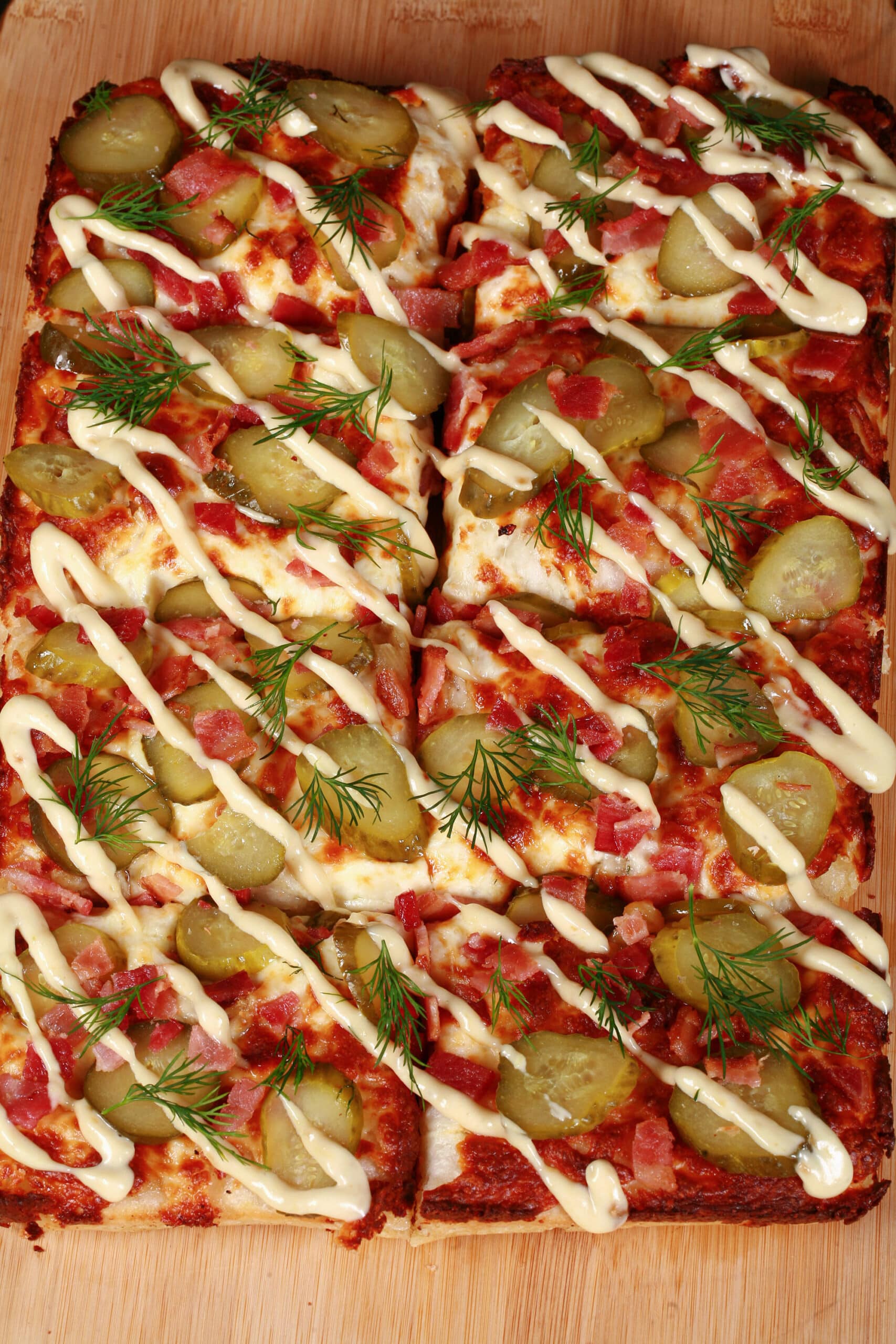 A detroit style gluten free pickle pizza, with dill pickle slices, bacon, fresh dill, and roasted garlic mayonnaise drizzled across it.