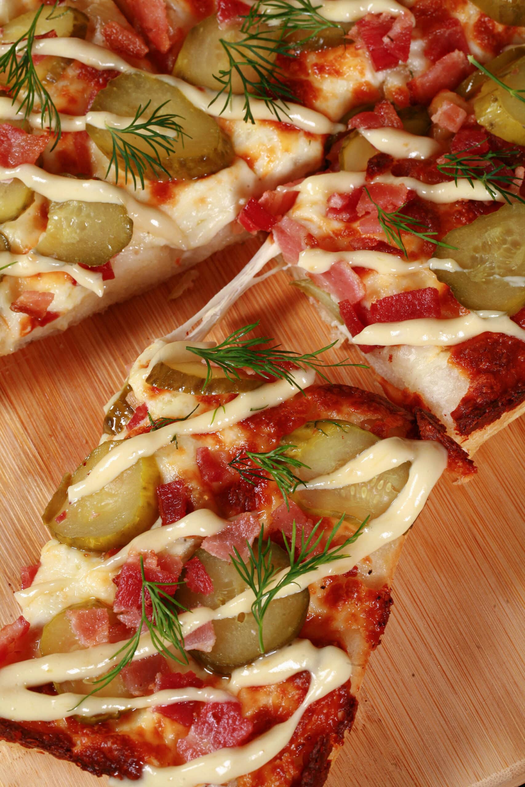 A detroit style gluten free pickle pizza, with dill pickle slices, bacon, fresh dill, and roasted garlic mayonnaise drizzled across it.