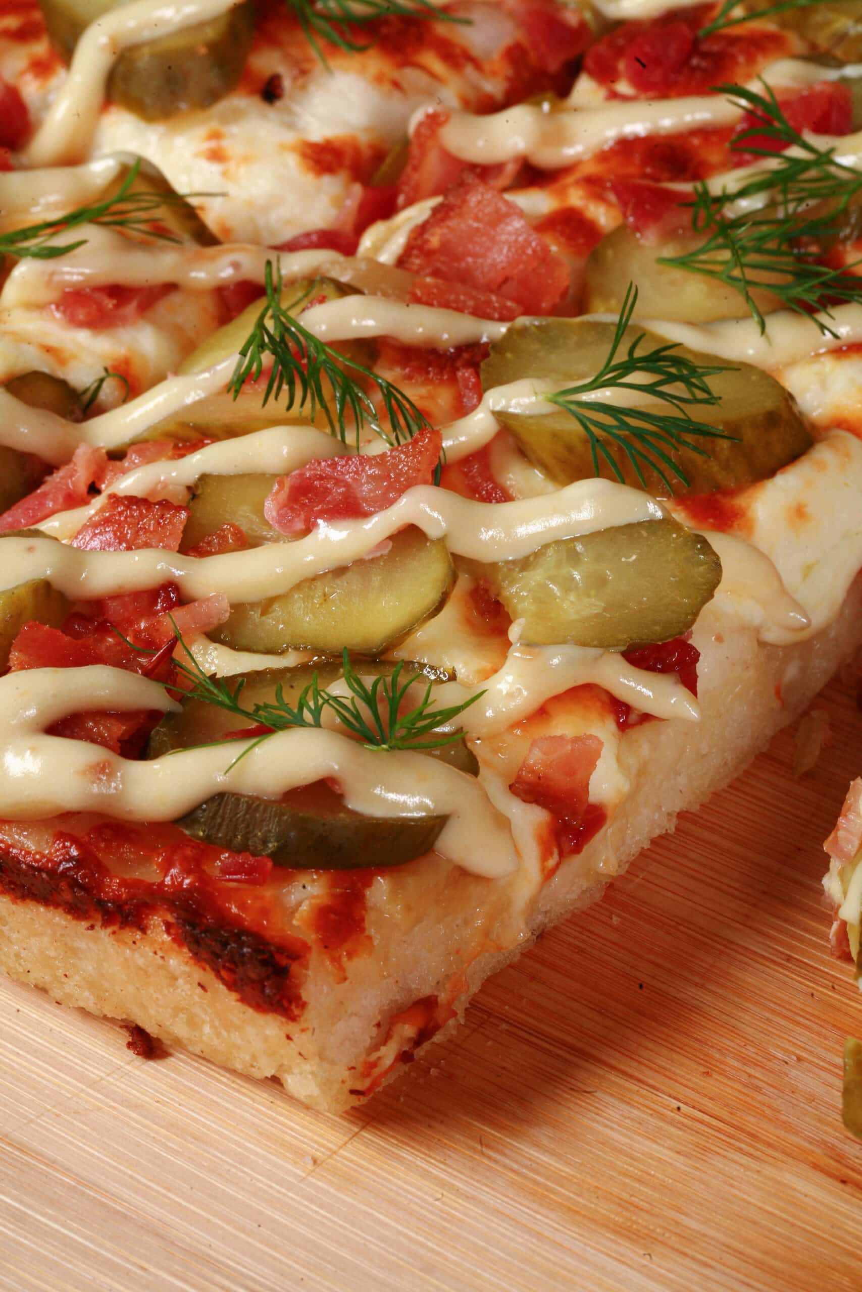 A detroit style gluten free dill pickle pizza, with pickle slices, bacon, fresh dill, and roasted garlic mayonnaise drizzled across it.