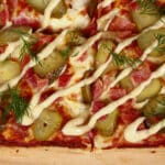 A detroit style gluten free pickle pizza, with dill pickle slices, bacon, fresh dill, and roasted garlic mayonnaise drizzled across it.