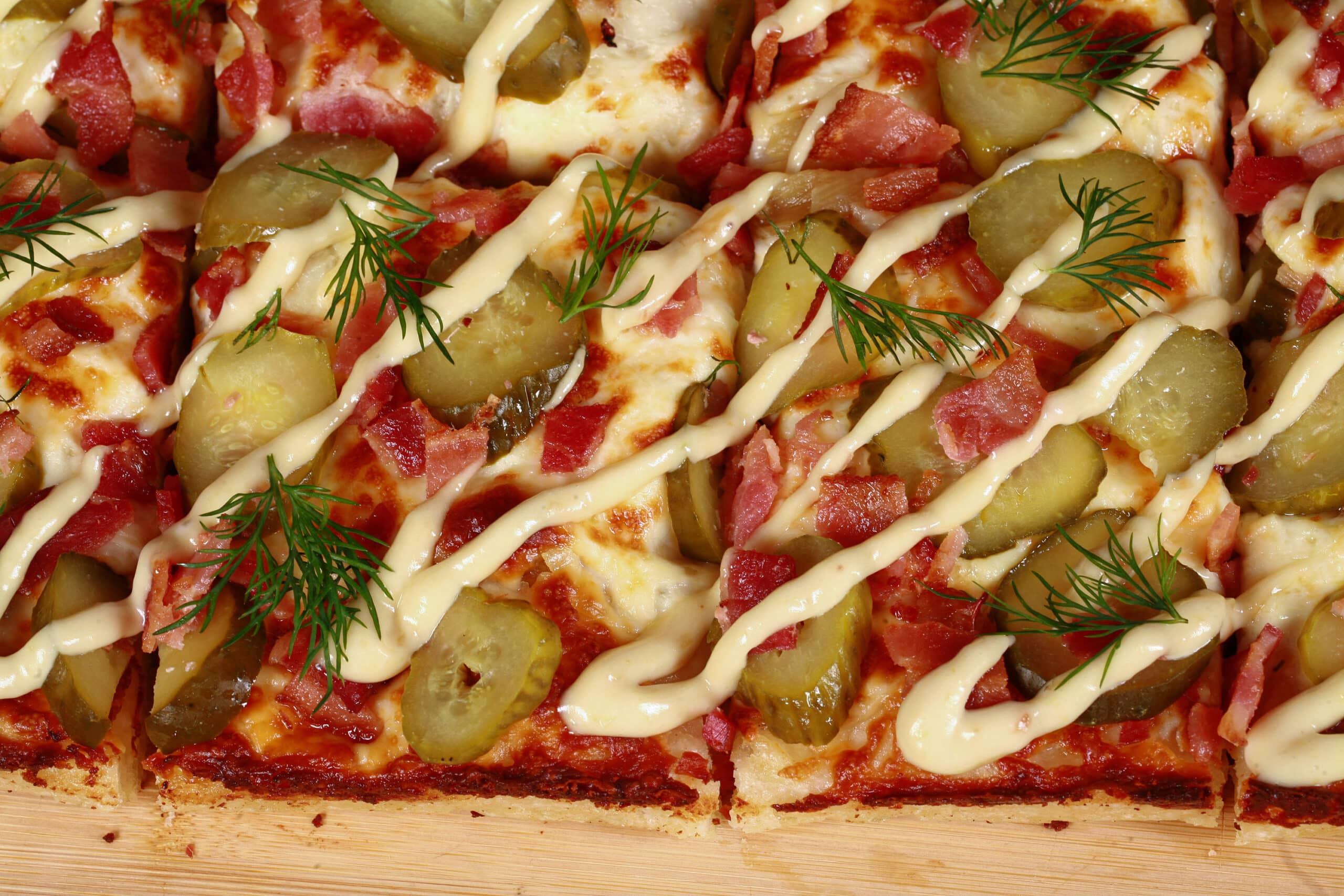 A detroit style gluten free pickle pizza, with dill pickle slices, bacon, fresh dill, and roasted garlic mayonnaise drizzled across it.