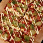 A detroit style gluten free dill pickle pizza, with pickle slices, bacon, fresh dill, and roasted garlic mayonnaise drizzled across it.