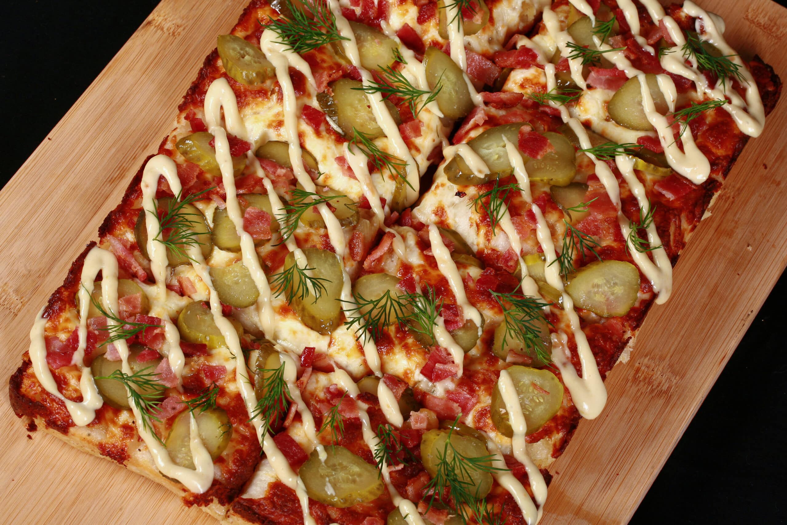 A detroit style gluten free dill pickle pizza, with pickle slices, bacon, fresh dill, and roasted garlic mayonnaise drizzled across it.